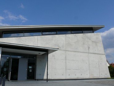 Ein modernes Gebäude mit Betonfassade und schrägem Dach vor blauem Himmel.