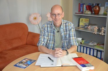 Ein Mann mit Brille und kariertem Hemd sitzt an einem Schreibtisch mit Büchern.