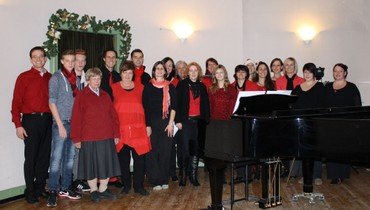 Ein Chor lächelnder Menschen, viele in Rot, stehen hinter einem Flügel.